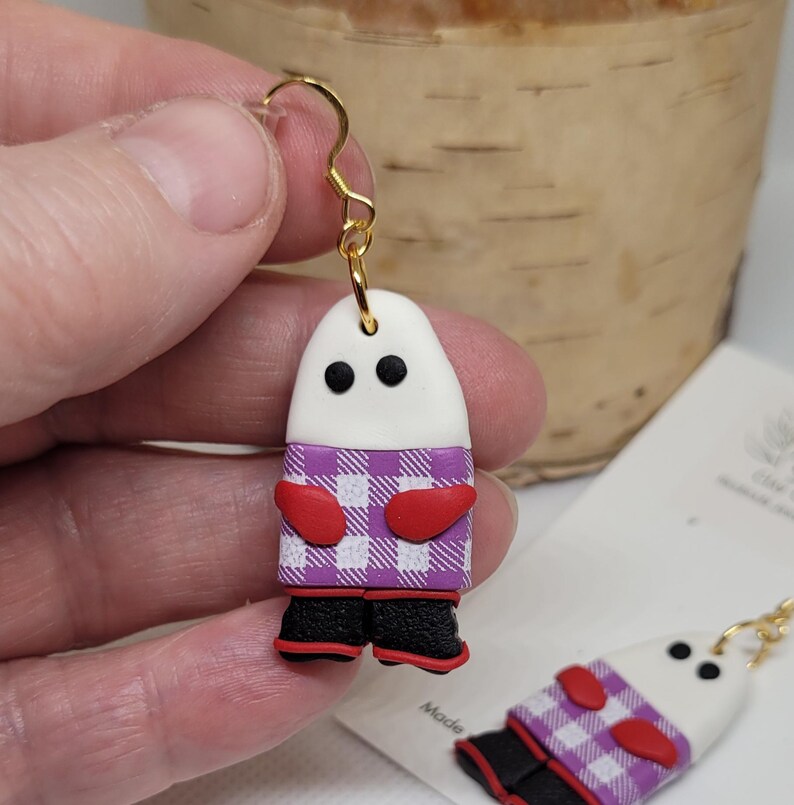 May include: A pair of white ghost-shaped earrings with purple and white checkered shirts, red mittens, and black boots. The earrings are made of polymer clay and have gold hooks.
