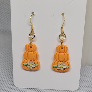 May include: A pair of orange pumpkin earrings with a white flower and green leaves. The earrings are made of polymer clay and have gold hooks.