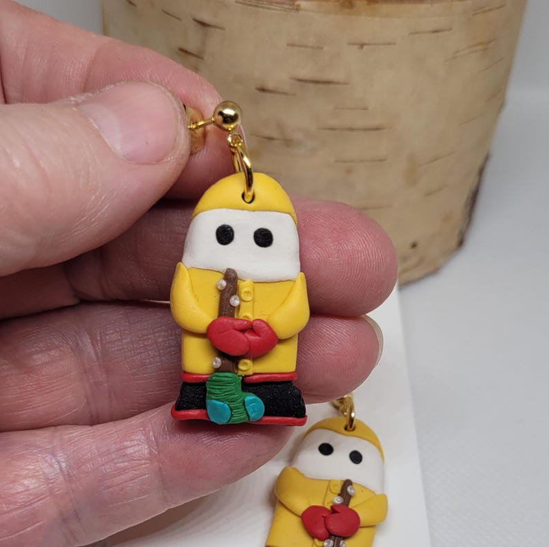May include: A pair of yellow polymer clay earrings shaped like a person wearing a yellow raincoat, red mittens, and blue boots. The person is holding a brown stick with white dots.