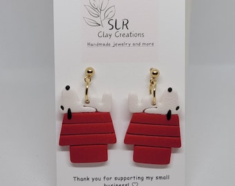 Puede incluir: Pendientes de arcilla roja y blanca con un perro de dibujos animados acostado encima de una caseta para perros. Los pendientes están en una tarjeta blanca con el texto "SLR Clay Creations Joyería hecha a mano y más" y "¡Gracias por apoyar mi pequeño negocio!"