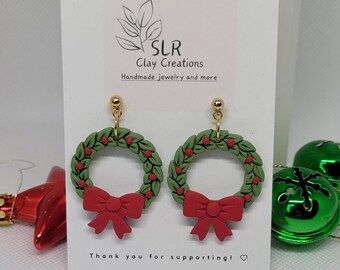 May include: A pair of handmade clay earrings featuring a Christmas wreath design. The wreaths are green with red berries and a red bow. The earrings are attached to gold-colored posts. The card behind the earrings reads "SLR Clay Creations" and "Thank you for supporting!"