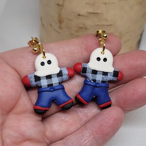 May include: A pair of dangle earrings featuring a ghost wearing a blue and white plaid shirt and blue jeans. The ghost has a white face with two black eyes. The earrings have gold hooks.
