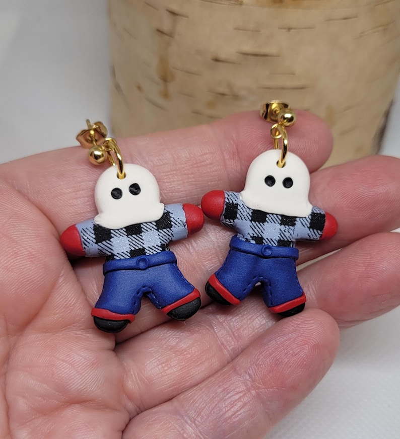 May include: A pair of dangle earrings featuring a ghost wearing a blue and white plaid shirt and blue jeans. The ghost has a white face with two black eyes. The earrings have gold hooks.