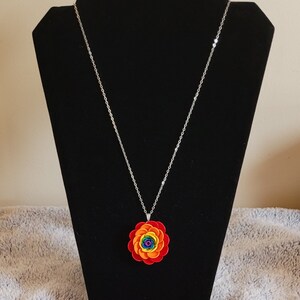 Peut inclure: Un collier en chaîne argentée avec un pendentif en forme de fleur arc-en-ciel. La fleur est composée de cercles de papier superposés en rouge, orange, jaune, vert, bleu, indigo et violet.