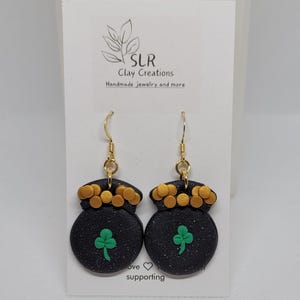 Peut inclure: Boucles d'oreilles pendantes noires et dorées avec un charme trèfle vert. Les boucles d'oreilles sont en forme de pot d'or avec un bord doré et des points dorés.