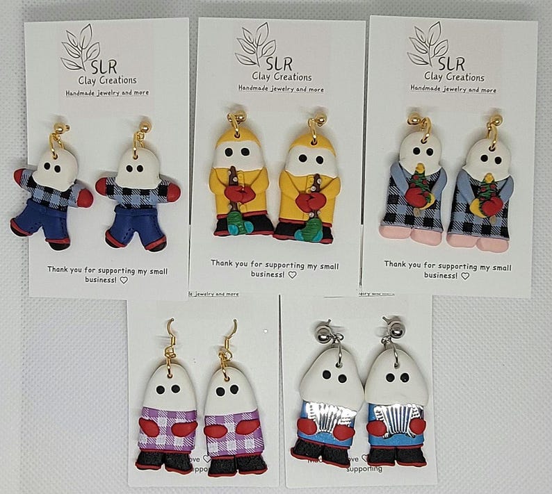 May include: Six pairs of handmade polymer clay earrings featuring a snowman design. Each pair features a different color scheme and pattern, including red, blue, yellow, white, and black. The earrings are all made with a white base and have a whimsical, cartoon-like style.