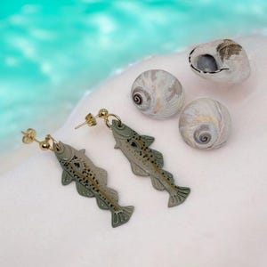 Peut inclure: Boucles d'oreilles dorées en forme de poisson avec des écailles et des nageoires détaillées. Les boucles d'oreilles sont présentées avec trois coquillages sur fond blanc, avec une eau turquoise floue en arrière-plan.