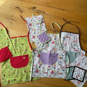 Könnte beinhalten: Vier farbenfrohe Küchenschürzen mit passenden Topflappen. Die Schürzen haben verschiedene Muster mit Gnomen, Füchsen und Bienen. Die Topflappen sind gesteppt und in verschiedenen Farben gehalten.