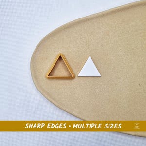 Op de afbeelding: Twee driehoekige objecten, één goudkleurig en één wit, worden weergegeven op een beige ovale ondergrond. De gouden driehoek is een frame, terwijl de witte massief is. De tekst "SHARP EDGES • MULTIPLE SIZES" staat onderaan.
