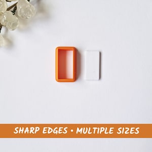 Puede incluir: Dos cortadores de galletas de plástico naranja, uno rectangular y otro cuadrado, sobre un fondo blanco. El texto "SHARP EDGES • MULTIPLE SIZES" está impreso en negro sobre una pancarta naranja en la parte inferior de la imagen.