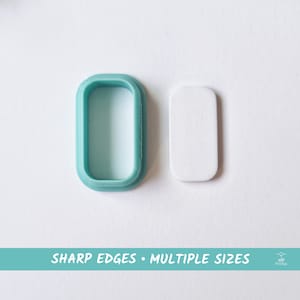 Peut inclure: Un emporte-pièce en plastique turquoise en forme de rectangle aux coins arrondis. À côté se trouve un rectangle blanc de fondant.