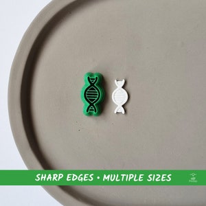 Könnte beinhalten: Zwei 3D-gedruckte DNA-Doppelhelix-Ausstecher, einer grün und einer weiß, auf einer grauen Oberfläche. Der Text "SHARP EDGES • MULTIPLE SIZES" ist unter den Ausstechern gedruckt.