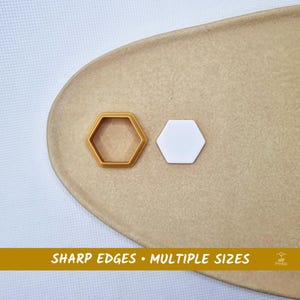 May include: Two hexagon-shaped cookie cutters, one gold and one white, are displayed on a tan surface. The gold cutter is an outline, while the white one is solid. Text at the bottom reads "SHARP EDGES • MULTIPLE SIZES."
