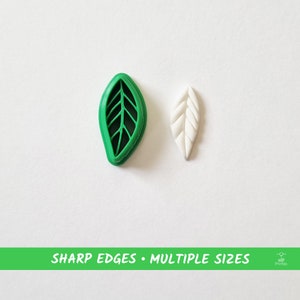 Puede incluir: Dos cortadores de galletas con forma de hoja, uno verde y otro blanco. El cortador verde tiene un patrón de vetas detallado. El cortador blanco es liso. El texto "SHARP EDGES • MULTIPLE SIZES" está impreso en una pancarta verde debajo de los cortadores.