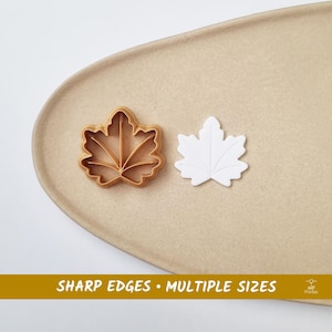 May include: A maple leaf cookie cutter and a white maple leaf shape are displayed on a beige surface. The cookie cutter is a light brown color. The text "SHARP EDGES • MULTIPLE SIZES" is in a gold bar at the bottom.