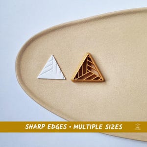 Op de afbeelding: Twee driehoekige objecten, één wit en één goudkleurig, liggen op een beige oppervlak. De witte driehoek heeft een gestructureerd, lineair ontwerp. De gouden driehoek heeft een geometrisch, opengewerkt patroon. De tekst onderaan luidt "SHARP EDGES • MULTIPLE SIZES."