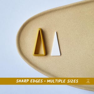 May include: Two triangular cookie cutters, one yellow and one white, are shown on a tan plate. The yellow cutter has a hollow centre, while the white one is solid. Text at the bottom reads "SHARP EDGES • MULTIPLE SIZES."
