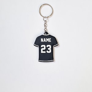 Può includere: Portachiavi a forma di maglia da calcio nero e bianco con il nome "NAME" e il numero "23" stampato sul retro.