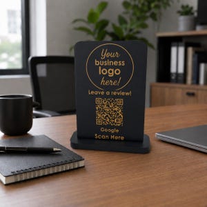Può includere: Un cartello rettangolare nero con base dorata che riporta il testo Your business logo here! Leave a review! e Google Scan Here, insieme a un codice QR centrale. Il cartello è progettato per l'esposizione aziendale e l'interazione.