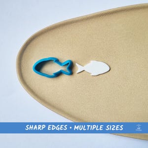 Puede incluir: Un cortador de galletas azul con forma de pez y un recorte blanco con forma de pez se muestran sobre una superficie beige. El texto "SHARP EDGES • MULTIPLE SIZES" está en la parte inferior de la imagen.