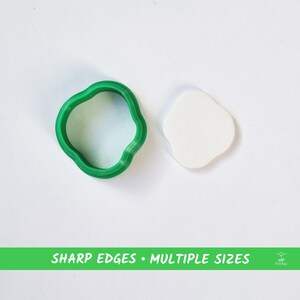 Pode incluir: Um cortador de biscoitos de plástico verde em forma de nuvem, ao lado de um biscoito branco em forma de nuvem. O texto "SHARP EDGES • MULTIPLE SIZES" está impresso em uma faixa verde abaixo da imagem.