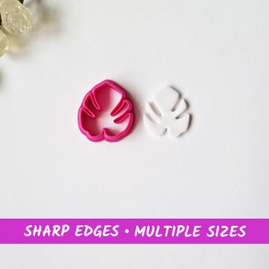 Puede incluir: Dos cortadores de galletas de plástico en forma de hoja de monstera. El cortador más grande es blanco y el más pequeño es rosa.