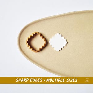 Può includere: Un tagliabiscotti a forma di diamante con bordo a zig-zag e una forma bianca sono su un piatto ovale beige. Il tagliabiscotti è marrone chiaro. Il testo "SHARP EDGES • MULTIPLE SIZES" è in basso.