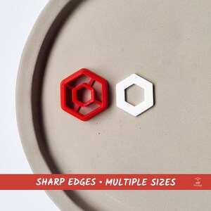 Puede incluir: Dos cortadores de galletas hexagonales impresos en 3D, uno rojo y otro blanco, sobre una superficie gris. El cortador rojo tiene un hexágono más pequeño en el interior. El texto "SHARP EDGES • MULTIPLE SIZES" está impreso debajo de los cortadores.