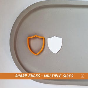 Op de afbeelding: Twee schildvormige koekjesuitstekers, een oranje en een wit, op een grijs oppervlak. De tekst "SHARP EDGES • MULTIPLE SIZES" is in oranje onderaan de afbeelding gedrukt.