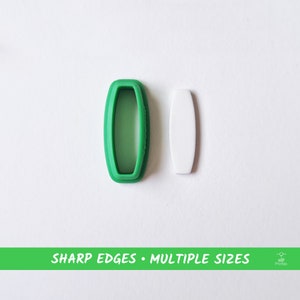 Puede incluir: Un cortador de galletas de plástico verde en forma de óvalo con un óvalo más pequeño cortado en el centro. Junto a él hay un óvalo de plástico blanco.