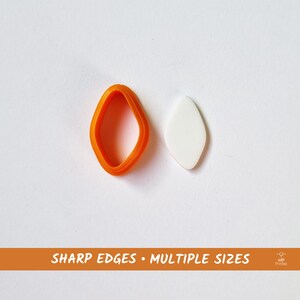 Puede incluir: Dos cortadores de galletas de plástico naranja en forma de diamante. Los cortadores están sobre una superficie blanca. El texto "SHARP EDGES • MULTIPLE SIZES" está impreso en una pancarta marrón debajo de los cortadores.