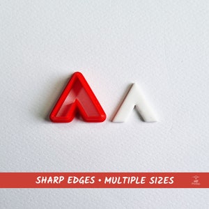 Puede incluir: Dos cortadores de galletas de plástico en forma de flecha. El cortador más grande es rojo y el más pequeño es blanco. El texto "SHARP EDGES - MULTIPLE SIZES" está impreso en rojo en la parte inferior de la imagen.