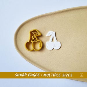 Op de afbeelding: Twee kersvormige koekjessnijders. Eén is geel, de andere is wit. De gele snijder ligt naast de witte snijder op een beige oppervlak. De tekst "SHARP EDGES • MULTIPLE SIZES" staat onderaan.