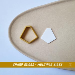 Pode incluir: Um cortador de biscoitos em forma geométrica dourada e uma forma geométrica branca são exibidos em uma superfície bege. O texto "SHARP EDGES • MULTIPLE SIZES" está na parte inferior da imagem.