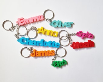 May include: A collection of eight colorful 3D-printed plastic keychains, each featuring a different name in a unique color: Emma in pink, Oliver in light blue, Anna in yellow, Charlotte in blue, James in orange, Elijah in green, Noah in red, and Sophia in magenta.