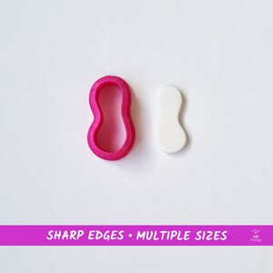 May include: A pink plastic cookie cutter shaped like a peanut, next to a white peanut-shaped cookie. The text "SHARP EDGES - MULTIPLE SIZES" is printed on a pink banner below the image.