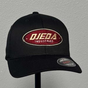 Peut inclure: Casquette de baseball noire ajustée avec un logo ovale brodé en rouge et or qui dit "OJEDA INDUSTRIES".