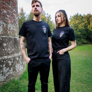 May include: Two people wearing black t-shirts featuring a skull design in a top hat with a pink bow tie. They are standing outdoors near a brick structure and green grass.