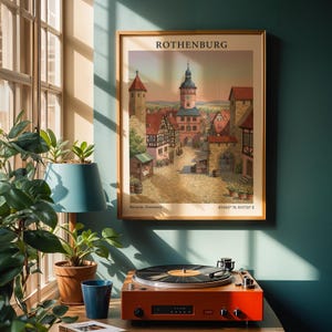 Op de afbeelding: Een ingelijste prent van Rothenburg, Duitsland, hangt aan een turquoise muur. De prent toont een kleurrijke illustratie van de architectuur van de stad. Een rode platenspeler staat op een houten oppervlak, met een blauwe lamp en planten in de buurt.