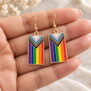 Puede incluir: Un par de pendientes rectangulares colgantes con marco de metal dorado e incrustaciones de esmalte de colores. El diseño muestra rayas verticales en morado, azul, verde, amarillo, naranja y rojo, rematadas con un patrón de chevrón en blanco, rosa y marrón.
