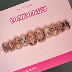 Puede incluir: Un conjunto de diez uñas postizas de color nude con diseños en blanco y negro. Los diseños incluyen un búho, gafas y las palabras "Gryffindor" y "Hogwarts". Las uñas son de Voodoo Nails.