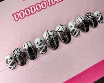 May include: A set of Halloween-themed press-on nails. The nails feature designs of spiders, spiderwebs, and mummy-like figures. The nails are black, white, and glittery. The text "Voodoo Nails" is visible on the pink background.