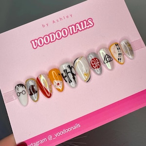 Puede incluir: Conjunto de diez uñas postizas blancas con varios diseños temáticos de Harry Potter, incluyendo el escudo de Hogwarts, el símbolo de las Reliquias de la Muerte y el número 9 3/4. Las uñas se muestran sobre un fondo rosa con el texto "VOODOO NAILS" e "Instagram @ _voodoonails".