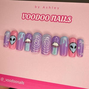 May include: A set of ten fake nails with a space theme. The nails are painted in shades of pink, purple, and blue. Some nails feature alien faces, while others have swirling patterns and a UFO. The text "VOODOO NAILS" is visible at the top of the image.
