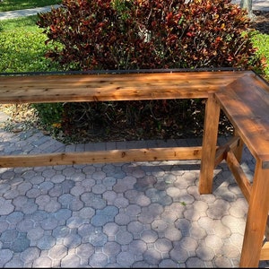 May include: An L-shaped wooden bar table featuring a polished brown finish, a long rectangular main section, and a shorter side extension. The table includes a thin black metal railing along the outer edges and dark metal corner brackets.