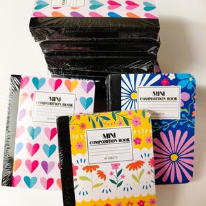 May include: A stack and arrangement of mini composition books, each wrapped in clear plastic. The covers feature colorful patterns including multi-colored hearts, blue floral designs, and yellow floral prints. Each book has a black spine and a white label with the text MINI COMPOSITION BOOK and 60 SHEETS.