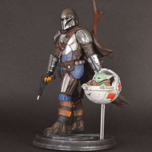 Op de afbeelding: Een zilveren en bruine Mandalorian action figure met een groen alien personage in een zwevende pod. De Mandalorian houdt een blastergeweer vast en heeft een cape. Het alien personage zit in de pod met zijn armen uitgestrekt.