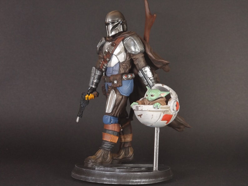 Op de afbeelding: Een zilveren en bruine Mandalorian action figure met een groen alien personage in een zwevende pod. De Mandalorian houdt een blastergeweer vast en heeft een cape. Het alien personage zit in de pod met zijn armen uitgestrekt.