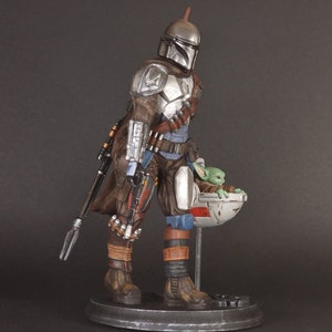 Op de afbeelding: Een zilveren en bruine Mandalorian action figure met een groen alien personage in een zwevende pod. De figuur houdt een wapen vast en draagt een helm.
