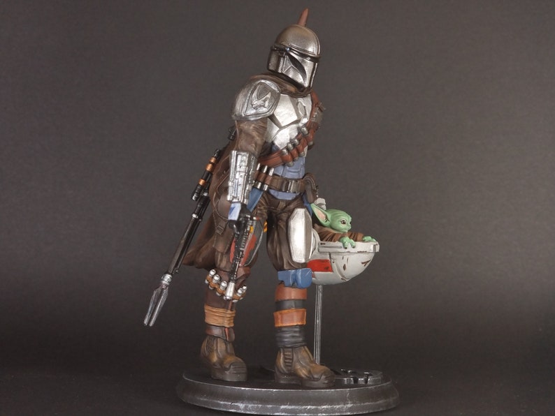 Op de afbeelding: Een zilveren en bruine Mandalorian action figure met een groen alien personage in een zwevende pod. De figuur houdt een wapen vast en draagt een helm.